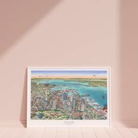 Hand-drawn Auckland map art print with digital colour, displayed in a white frame. Features Skytower, CBD, waterfront, docks, bridge, and America's Cup boats. New Zealand.